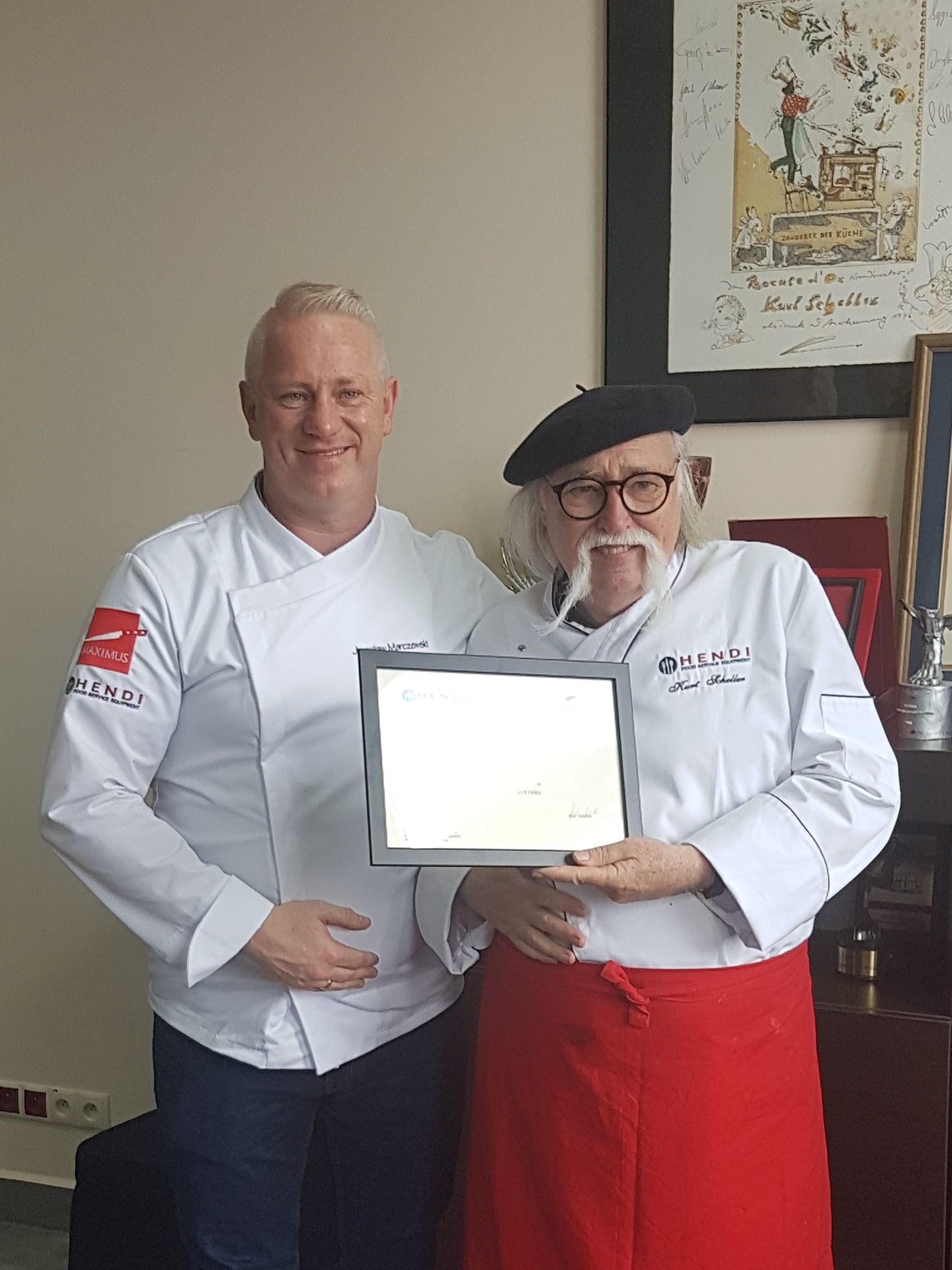 Dwóch kucharzy w białych uniformach prezentuje certyfikat firmy Hendi, jeden w czarnym berecie i czerwonym fartuchu, w tle rysunek z motywem kulinarnym.