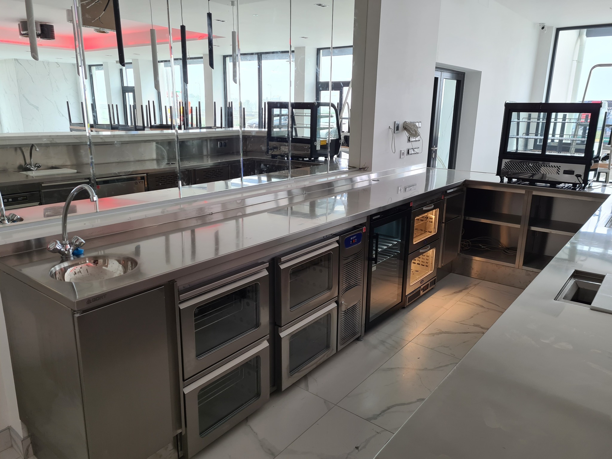 Profesjonalne wyposażenie gastronomiczne ze stali nierdzewnej: zlewozmywak, piekarniki konwekcyjne i chłodziarka, w jasnym wnętrzu restauracji.