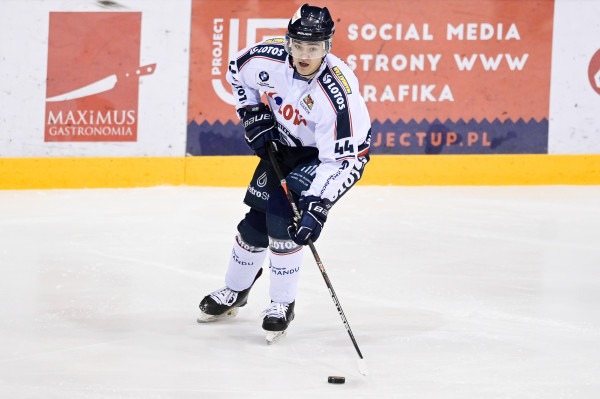 Hokeista w akcji na lodowisku, w tle widoczne reklamy, w tym logo Maximus Gastronomia.