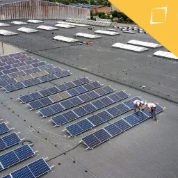 Widok z góry na instalację paneli słonecznych na płaskim dachu budynku przemysłowego; dwóch techników w białych kaskach i kombinezonach łączy panele, widoczne kable i logo firmy w prawym górnym rogu.