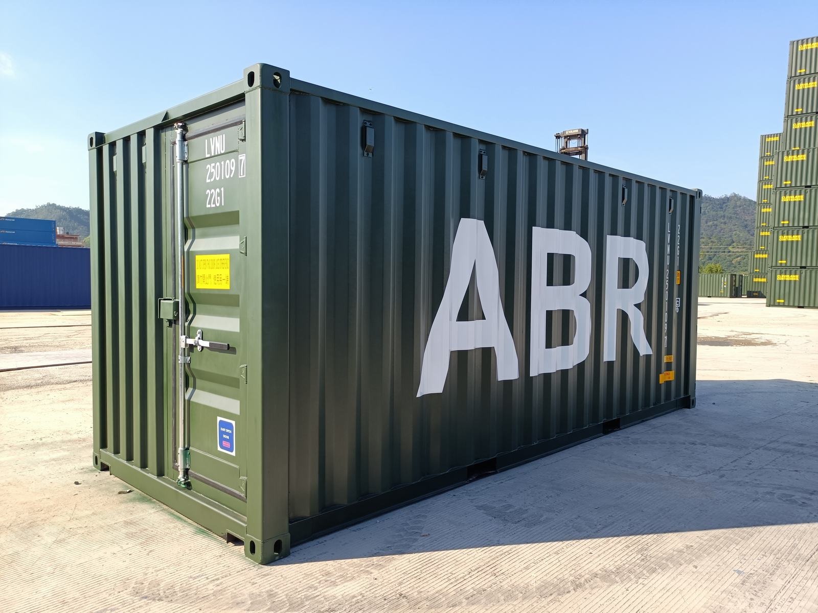Nowy, oliwkowozielony kontener transportowy z białym napisem ABR, stojący na betonowym placu, w tle góra i inne kontenery.