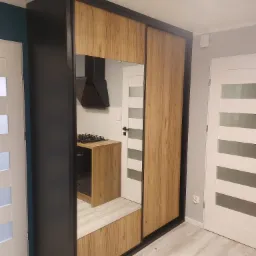 Nowoczesna szafa przesuwna z lustrem, w której odbija się fragment kuchni z drewnianym blatem i czarnym okapem, umieszczona między dwoma białymi drzwiami z przeszkleniami.