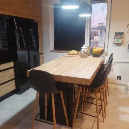 Wysoki stół z blatem imitującym drewno, otoczony czarnymi krzesłami barowymi z drewnianymi nogami, w tle zabudowa kuchenna z frontami w kolorze drewna i czarny sprzęt AGD.