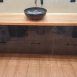 Łazienkowa szafka pod umywalkę z blatem z jasnego drewna i czarnymi, lakierowanymi frontami szuflad, umywalka w kształcie misy, widoczne płytki podłogowe imitujące drewno.