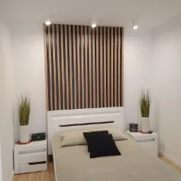 Nowoczesna sypialnia z białym łóżkiem, drewnianym panelem dekoracyjnym za wezgłowiem, białymi szafkami nocnymi i roślinami w doniczkach.
