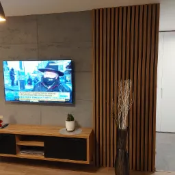 Nowoczesny salon z betonową ścianą, drewnianą lamelową ścianką działową, telewizorem, szafką RTV z czarnymi frontami, dekoracjami świątecznymi i wazonem z suchymi gałązkami