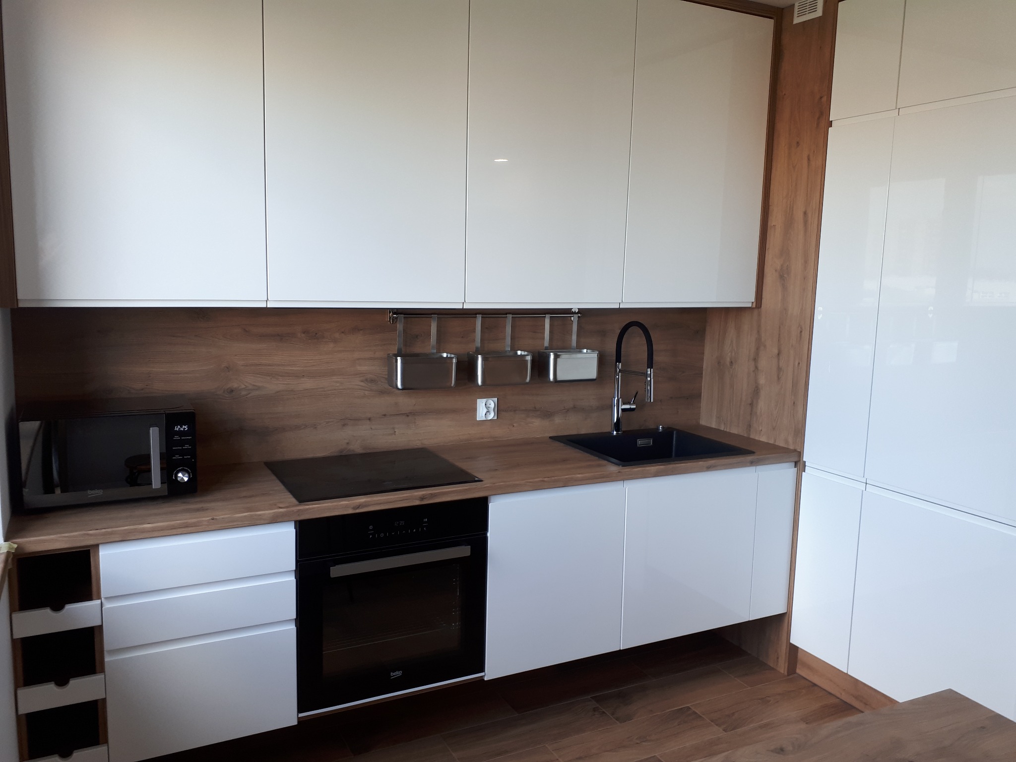 Nowoczesna kuchnia z białymi, błyszczącymi frontami szafek i drewnianym blatem, wyposażona w czarny zlew, płytę indukcyjną i piekarnik marki Beko, z widocznym mikrofalówką i relingiem na akcesoria...