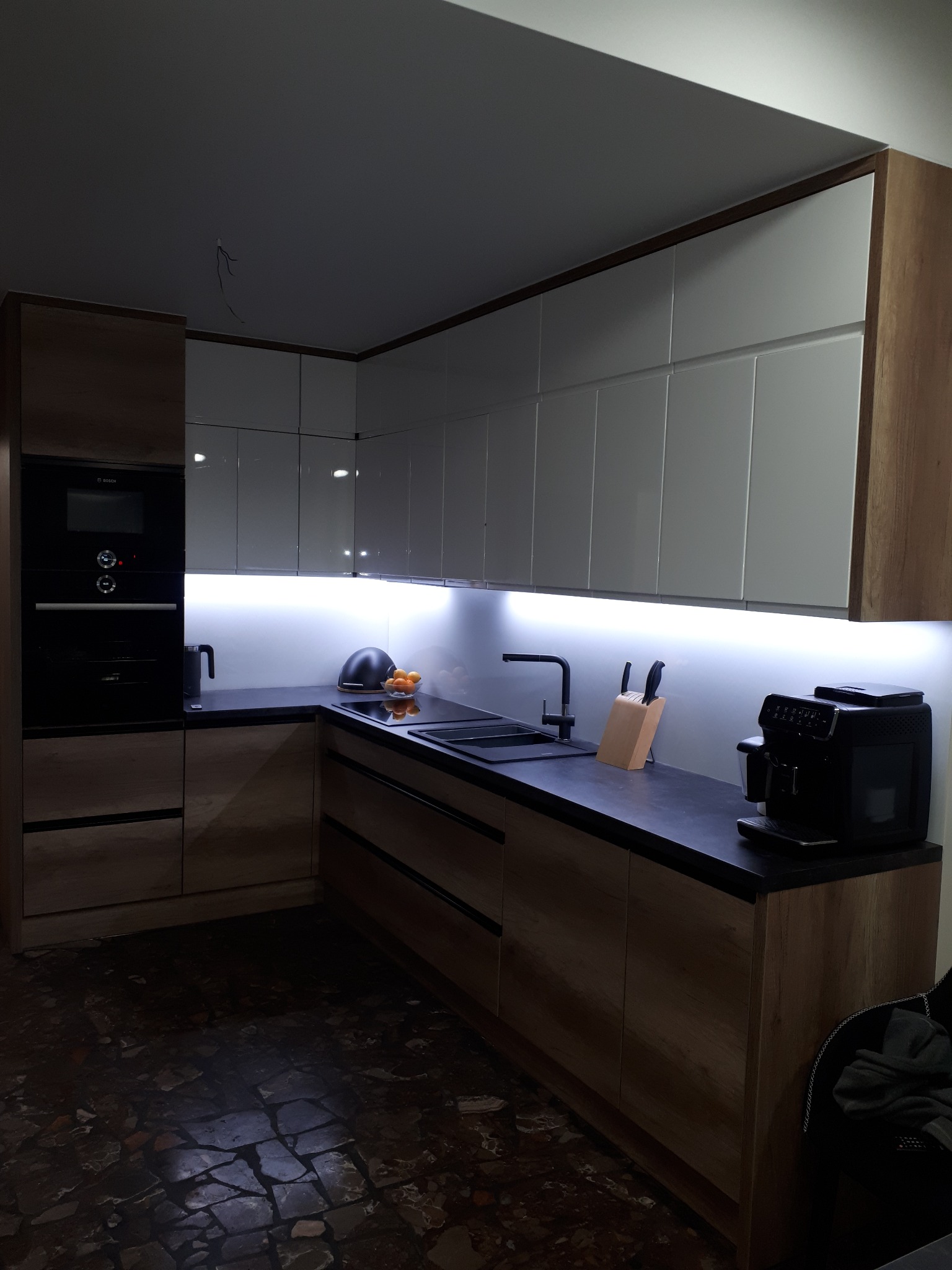 Nowoczesna kuchnia z białymi górnymi szafkami i drewnopodobnymi dolnymi, czarnym blatem, oświetleniem LED podszafkowym i czarną armaturą, widoczny ekspres do kawy i owoce w misce.