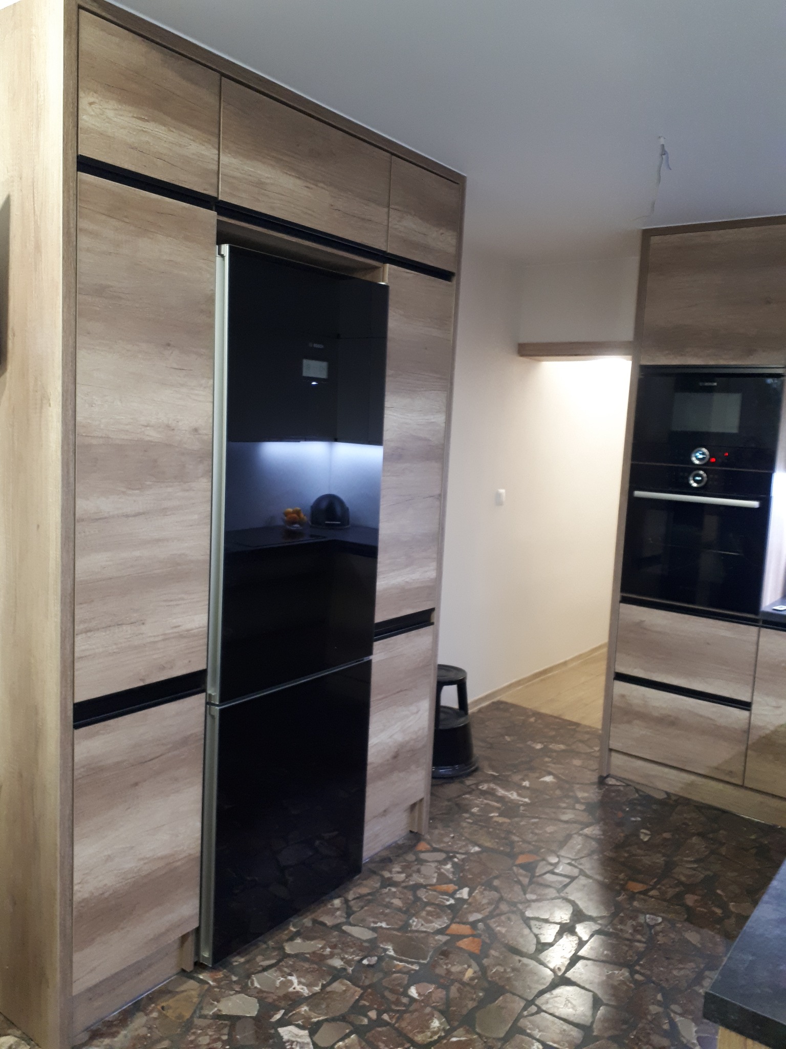 Nowoczesna kuchnia z zabudową w odcieniach drewna i czarnymi akcentami, z czarną lodówką w zabudowie, widok na fragment wnętrza z mozaikową podłogą.