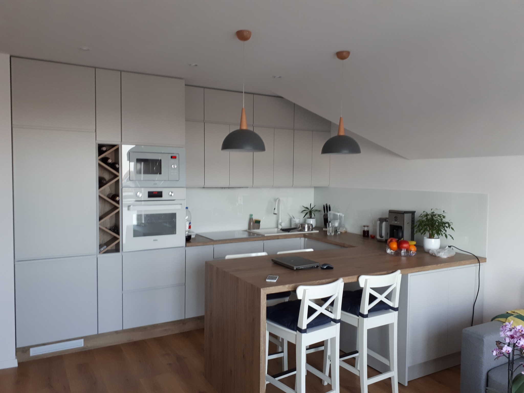 Jasna kuchnia z drewnianym blatem, białymi krzesłami barowymi i nowoczesnymi lampami wiszącymi, laptop na blacie, widok na skośny sufit.