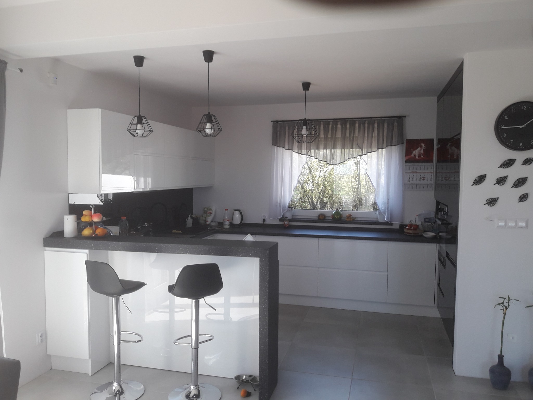 Nowoczesna, jasna kuchnia z białymi frontami i czarnym blatem, oświetlona trzema lampami wiszącymi w stylu industrialnym, z dwoma hokerami przy wyspie i widokiem na ogród przez okno.