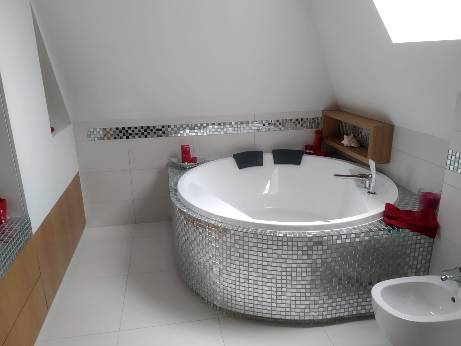 Nowoczesna łazienka na poddaszu z okrągłą wanną obłożoną mozaiką lustrzaną, białą armaturą i akcentami dekoracyjnymi w odcieniach czerwieni.
