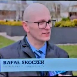 SEKSUOLOG TERAPEUTA PSYCHOLOG - Łysy mężczyzna w okularach, garniturze i niebieskim krawacie, mówiący na zewnątrz. Na dole ekranu pasek z nazwiskiem 'RAFAŁ SKOCZEK', psycholog i terapeuta. Tło to zieleń i donice z żółtymi krzewami.