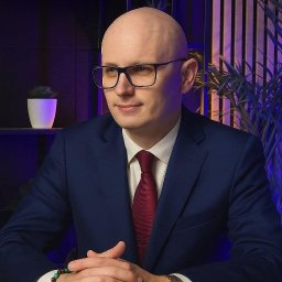 SEKSUOLOG TERAPEUTA PSYCHOLOG - Portret łysiejącego mężczyzny w okularach, granatowym garniturze i czerwonym krawacie, z dłońmi splecionymi na stole. Tło z pionowymi listwami i rośliną, oświetlone fioletowo-niebieskim światłem.