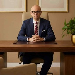 SEKSUOLOG TERAPEUTA PSYCHOLOG - Łysy mężczyzna w okularach, granatowym garniturze i krawacie, siedzi za drewnianym biurkiem ze splecionymi dłońmi. W tle beżowa ściana z obrazami i rośliną.