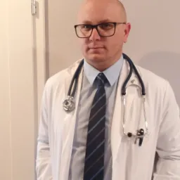 Portret lekarza w okularach i białym fartuchu ze stetoskopem, stojącego na tle jasnej ściany i białych drzwi.