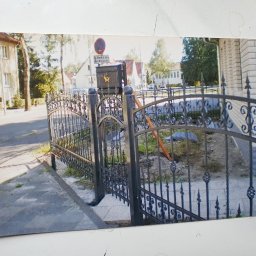 BAK-STAL - Kute, czarne ogrodzenie z dekoracyjnymi elementami, widoczne z zewnątrz posesji, z ulicą i budynkami w tle, w trakcie montażu.