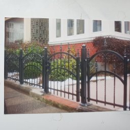 BAK-STAL - Czarne, metalowe ogrodzenie z dekoracyjnymi łukami i pionowymi prętami, zamontowane na niskim, betonowym murku przed domem z ceglaną elewacją i zielenią w tle.