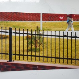 BAK-STAL - Czarne, metalowe ogrodzenie z ceglaną podmurówką, na tle trawnika i ceglanego budynku. W tle osoba z dzieckiem przechodząca obok znaku drogowego z symbolem psa.