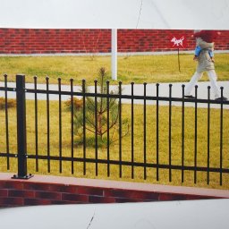 BAK-STAL - Zdjęcie czarnego metalowego ogrodzenia na niskiej ceglanej podmurówce, z osobą trzymającą dziecko przechodzącą w tle, obok trawnika i budynku z czerwonej cegły.