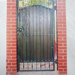 BAK-STAL - Drewniana brama ogrodzeniowa z metalowymi zdobieniami, osadzona między ceglanymi słupkami, widoczny detal dekoracyjny w dolnej części bramy.