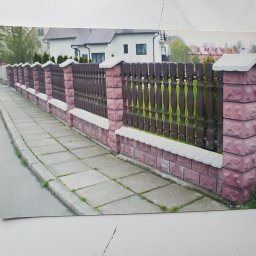 BAK-STAL - Długie, ozdobne ogrodzenie z brązowych, drewnianych sztachet i różowych, kamiennych słupków z białymi nakrywami, biegnące wzdłuż chodnika z szarych płyt.