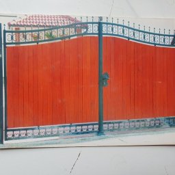 BAK-STAL - Zdjęcie przedstawia zamkniętą, drewnianą bramę w kolorze ceglastym, z czarnymi, ozdobnymi elementami metalowymi u góry i u dołu. Brama jest sfotografowana na tle fragmentu domu z czerwonym dachem.