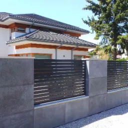 Nowoczesne ogrodzenie z poziomych, ciemnych paneli i betonowych słupków przed domem z białymi ścianami i ciemną dachówką.