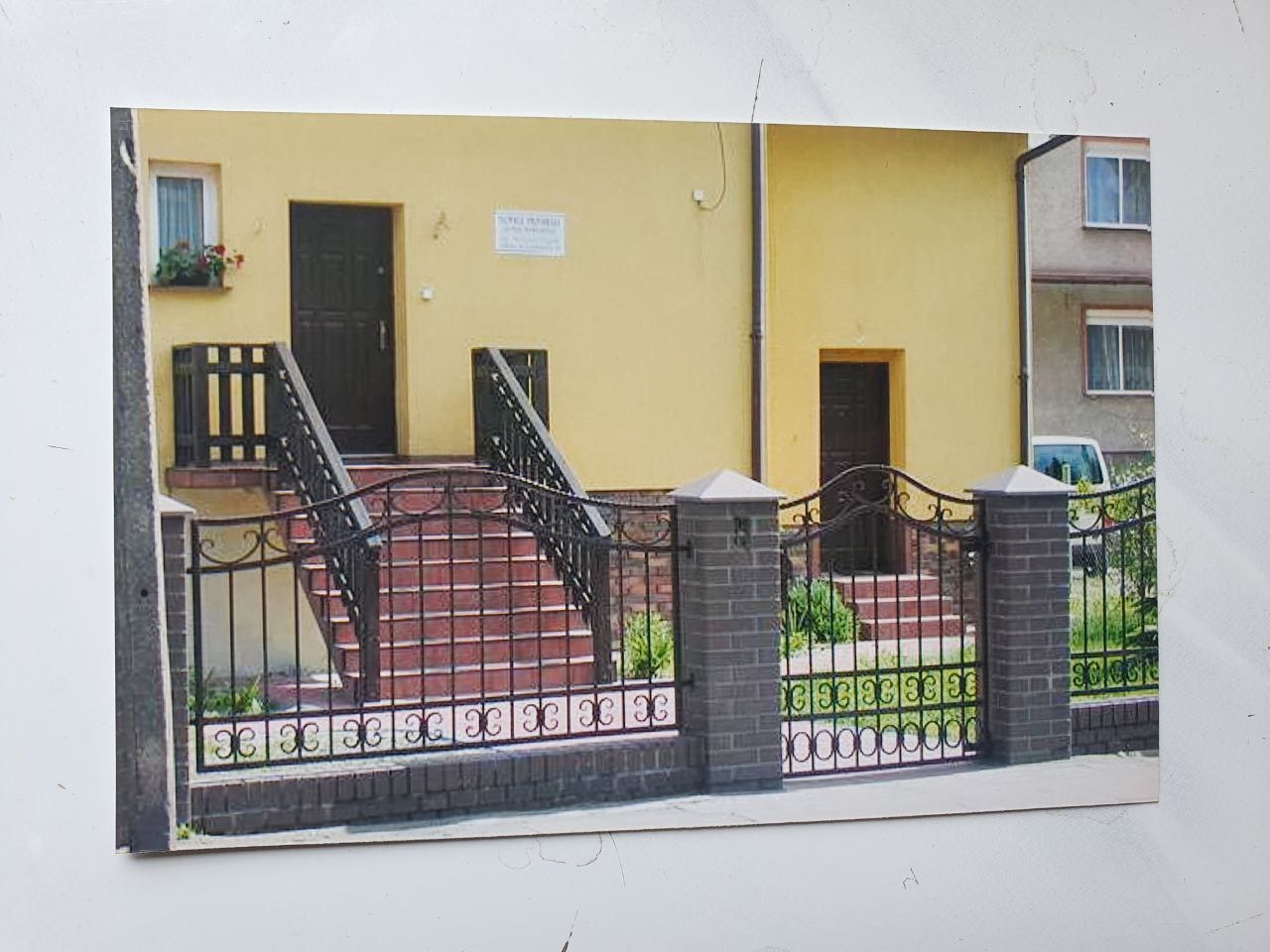 Kuta, czarna balustrada i ogrodzenie z ceglanymi słupkami przed żółtym budynkiem z dwoma wejściami i schodami wyłożonymi czerwoną cegłą klinkierową. Elementy dekoracyjne z motywem roślinnym.