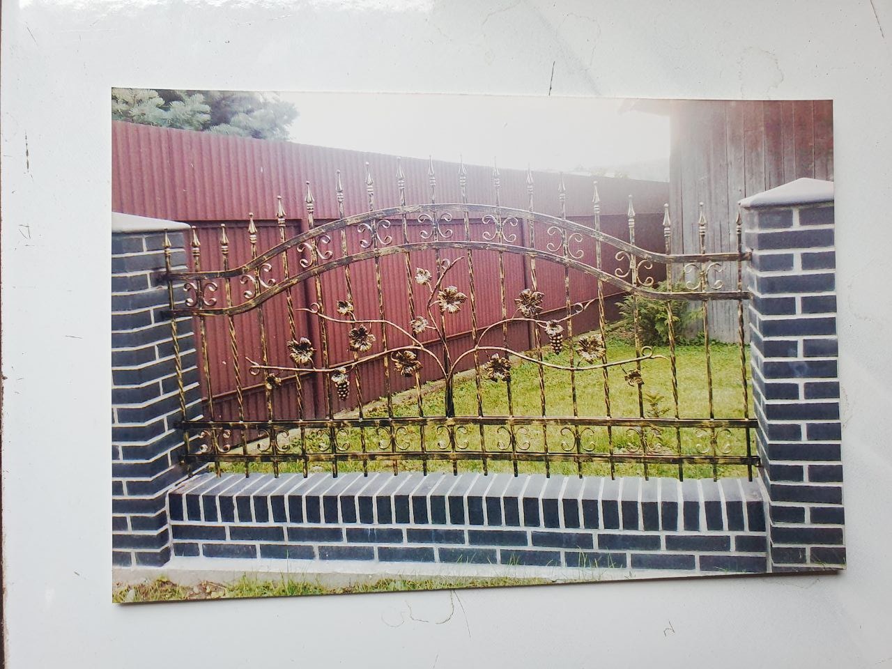 Kute, zdobione motywem winorośli ogrodzenie w Krakowie, osadzone na niskim murku z cegły klinkierowej, z tłem w postaci brązowego ogrodzenia panelowego i zieleni.