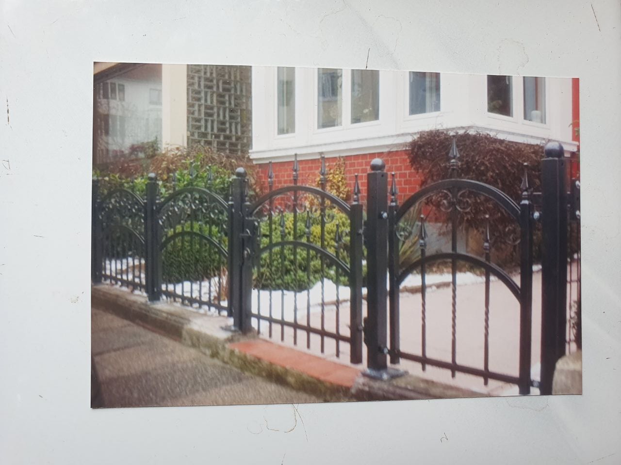 Czarne, metalowe ogrodzenie z dekoracyjnymi łukami i pionowymi prętami, zamontowane na niskim, betonowym murku przed domem z ceglaną elewacją i zielenią w tle.