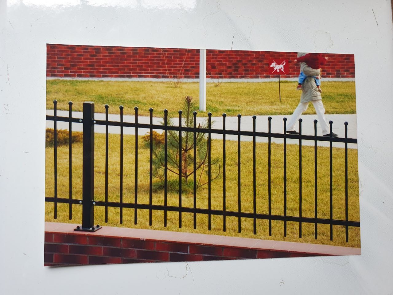 Czarne, metalowe ogrodzenie z ceglaną podmurówką, na tle trawnika i ceglanego budynku. W tle osoba z dzieckiem przechodząca obok znaku drogowego z symbolem psa.