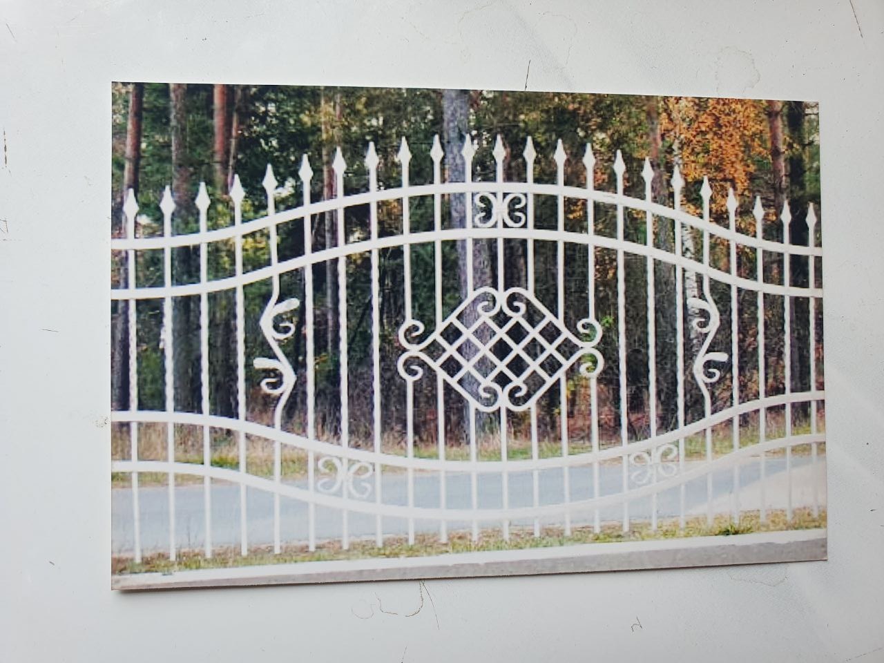 Zdjęcie białego, metalowego ogrodzenia z ozdobnymi elementami na tle lasu. Widoczne proste, pionowe pręty oraz poziome elementy dekoracyjne w kształcie fal i rombów. Ogrodzenie pomalowane na biało,...