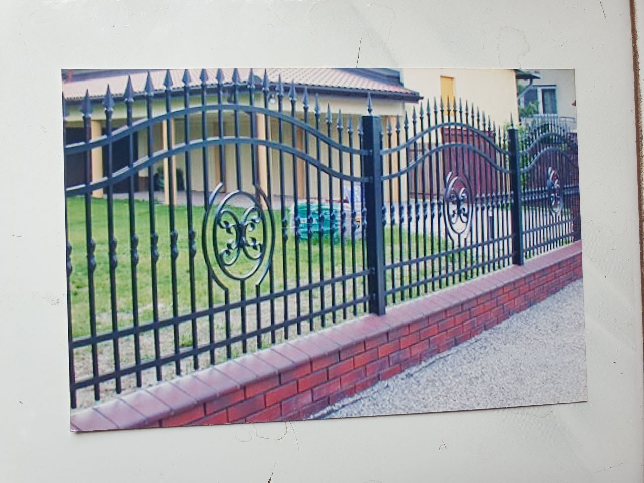 Czarne, metalowe ogrodzenie z dekoracyjnymi elementami na ceglanej podmurówce, widoczne z boku, z domem w tle.