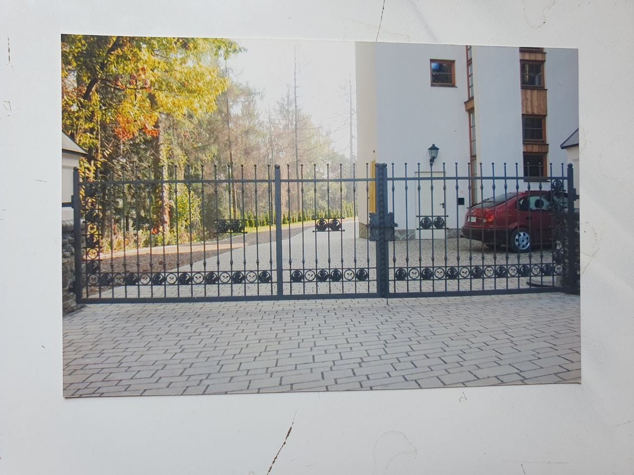 Czarna, metalowa brama wjazdowa z dekoracyjnymi elementami, prowadząca do posesji z białym budynkiem i czerwonym samochodem w tle, sfotografowana na tle brukowanej nawierzchni.