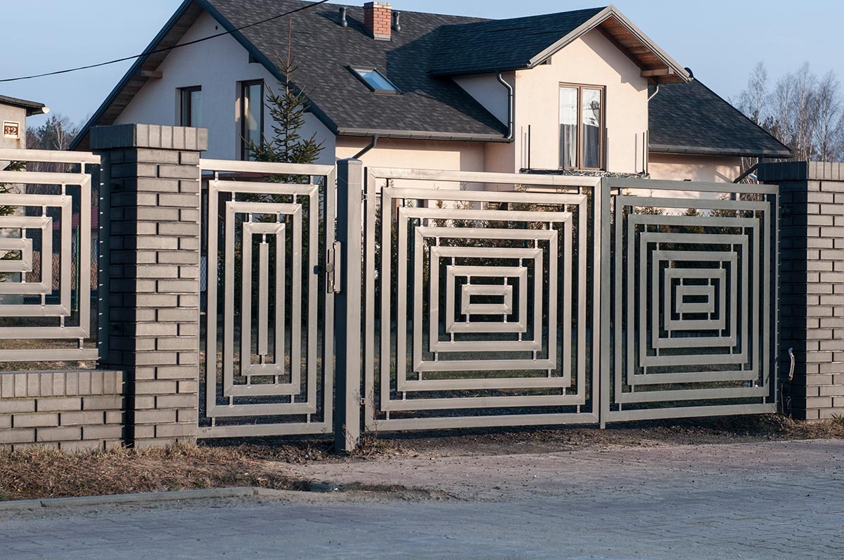 Nowoczesna, metalowa brama wjazdowa z geometrycznym wzorem kwadratów, osadzona na słupkach z szarej cegły klinkierowej, na tle domu z ciemnym dachem.