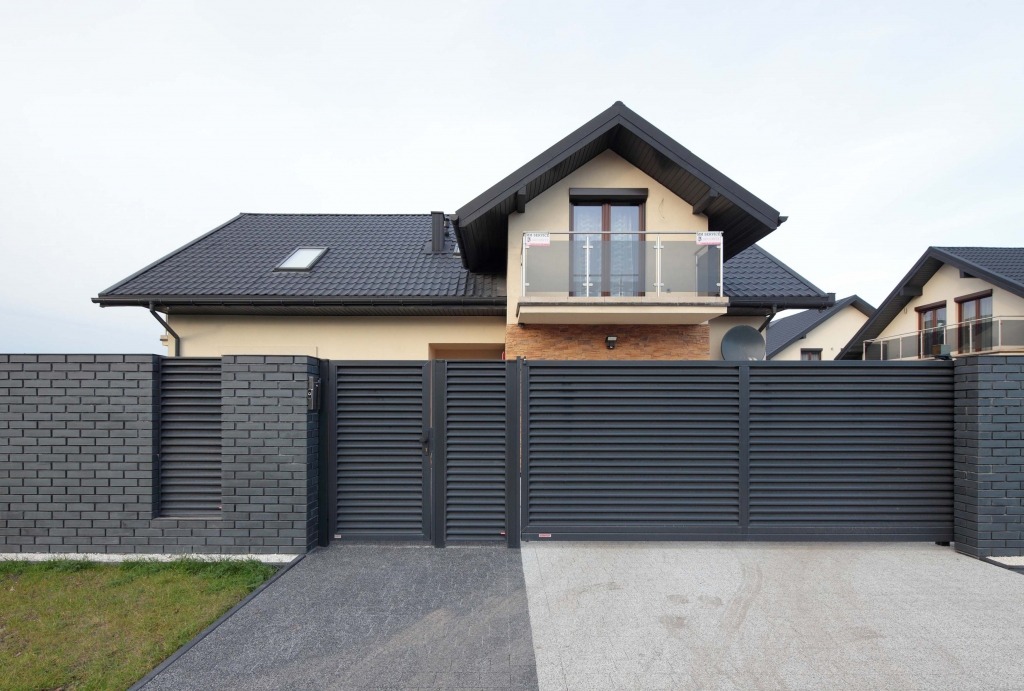 Nowoczesna posesja z przesuwną bramą lamelową w kolorze antracytowym, zintegrowaną z murem z szarej cegły i podjazdem o dwóch różnych fakturach, na tle domu z balkonem z balustradą szklaną.