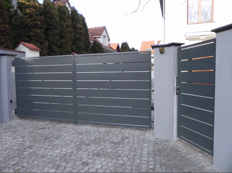 Nowoczesna, grafitowa brama wjazdowa i furtka z poziomych profili, zamontowane na murowanych słupkach, na tle kostki brukowej i zabudowy jednorodzinnej.