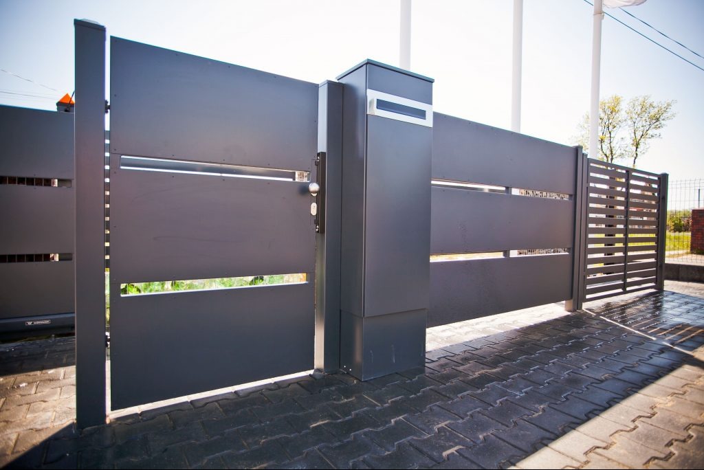 Nowoczesna, przesuwna brama wjazdowa z poziomymi szczelinami, pomalowana na ciemny szary kolor, z widocznym mechanizmem otwierania i skrzynką na listy, na tle kostki brukowej.