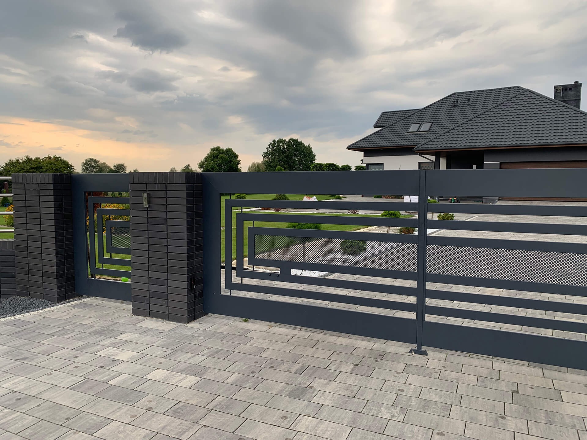 Nowoczesna brama wjazdowa z szarej stali z geometrycznym wzorem, osadzona na murowanych słupkach z cegły klinkierowej, na tle domu z ciemną dachówką i pochmurnego nieba.