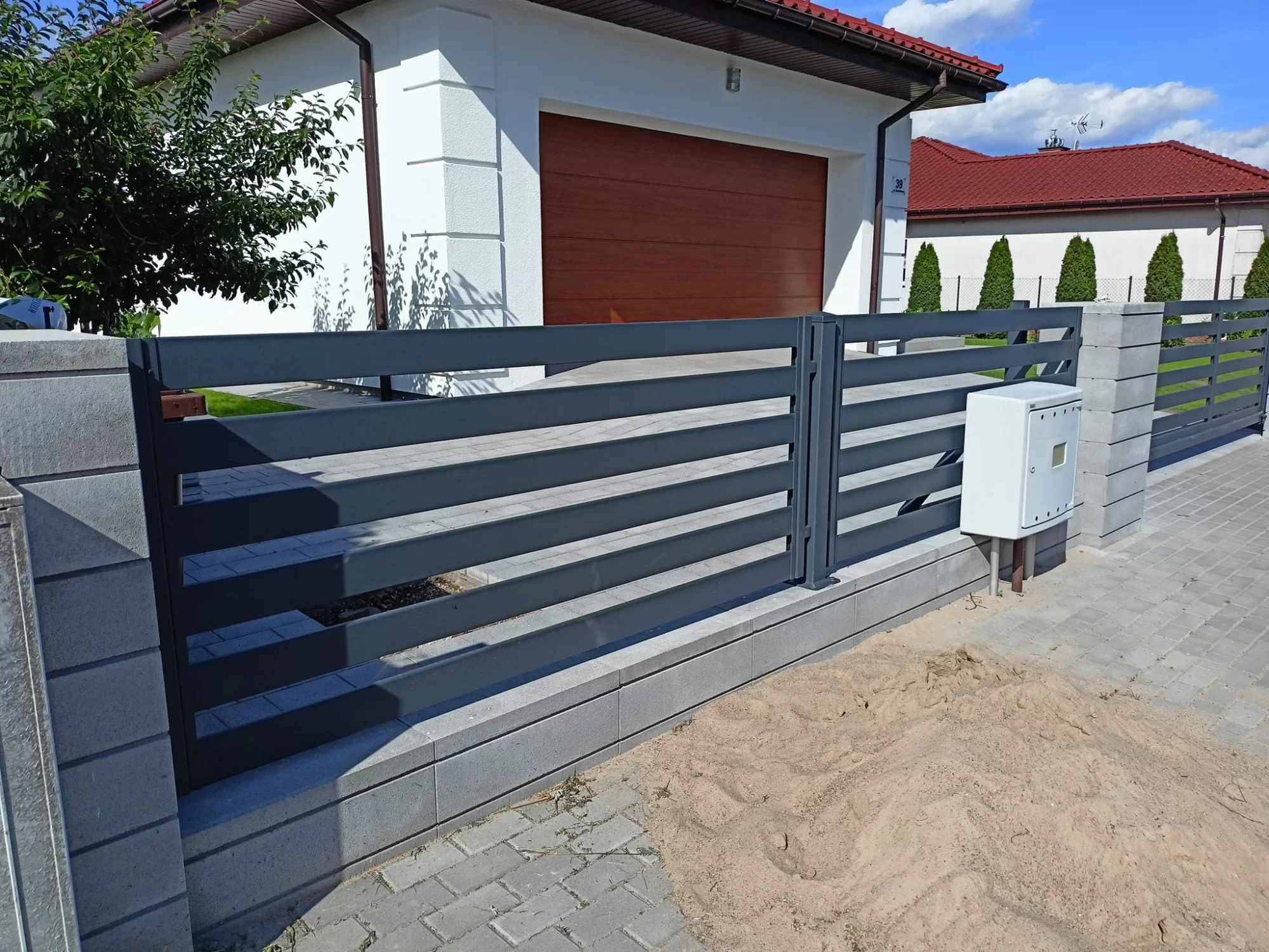 Nowoczesne, poziome przęsła ogrodzeniowe w kolorze grafitowym, osadzone na betonowych słupkach i podmurówce, z bramką wejściową i skrzynką pocztową, na tle jasnego domu z brązową bramą garażową.