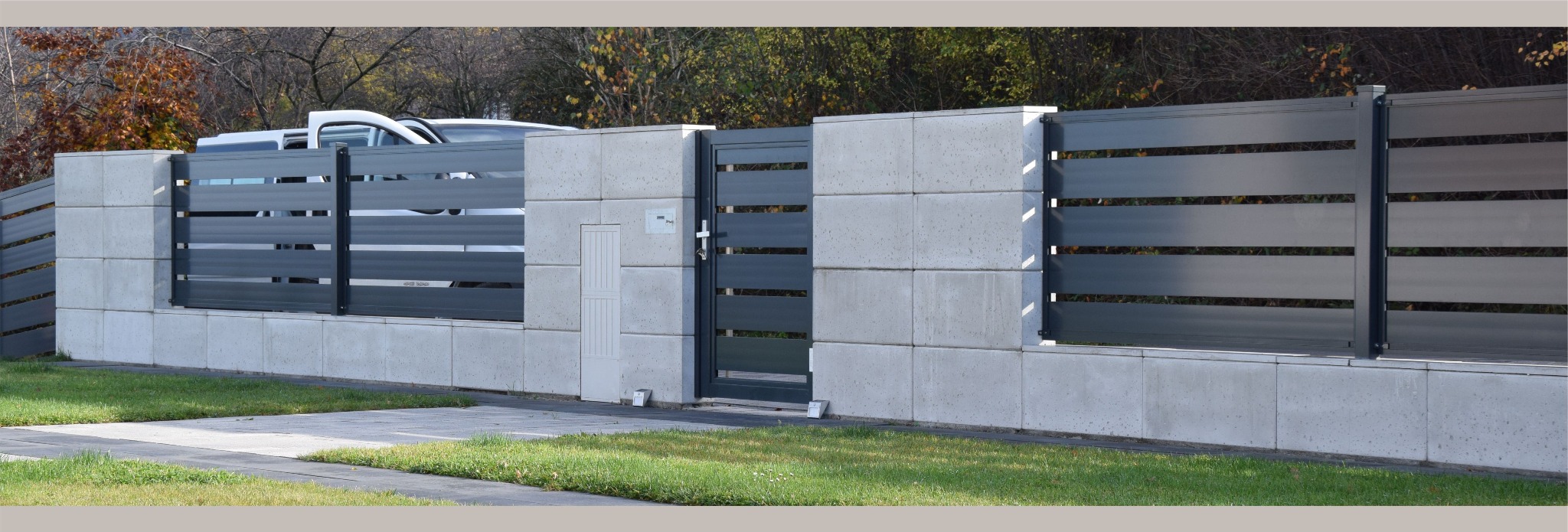 Nowoczesne ogrodzenie z betonowych bloczków i poziomych, szarych paneli, z furtką wejściową i widocznym samochodem w tle.