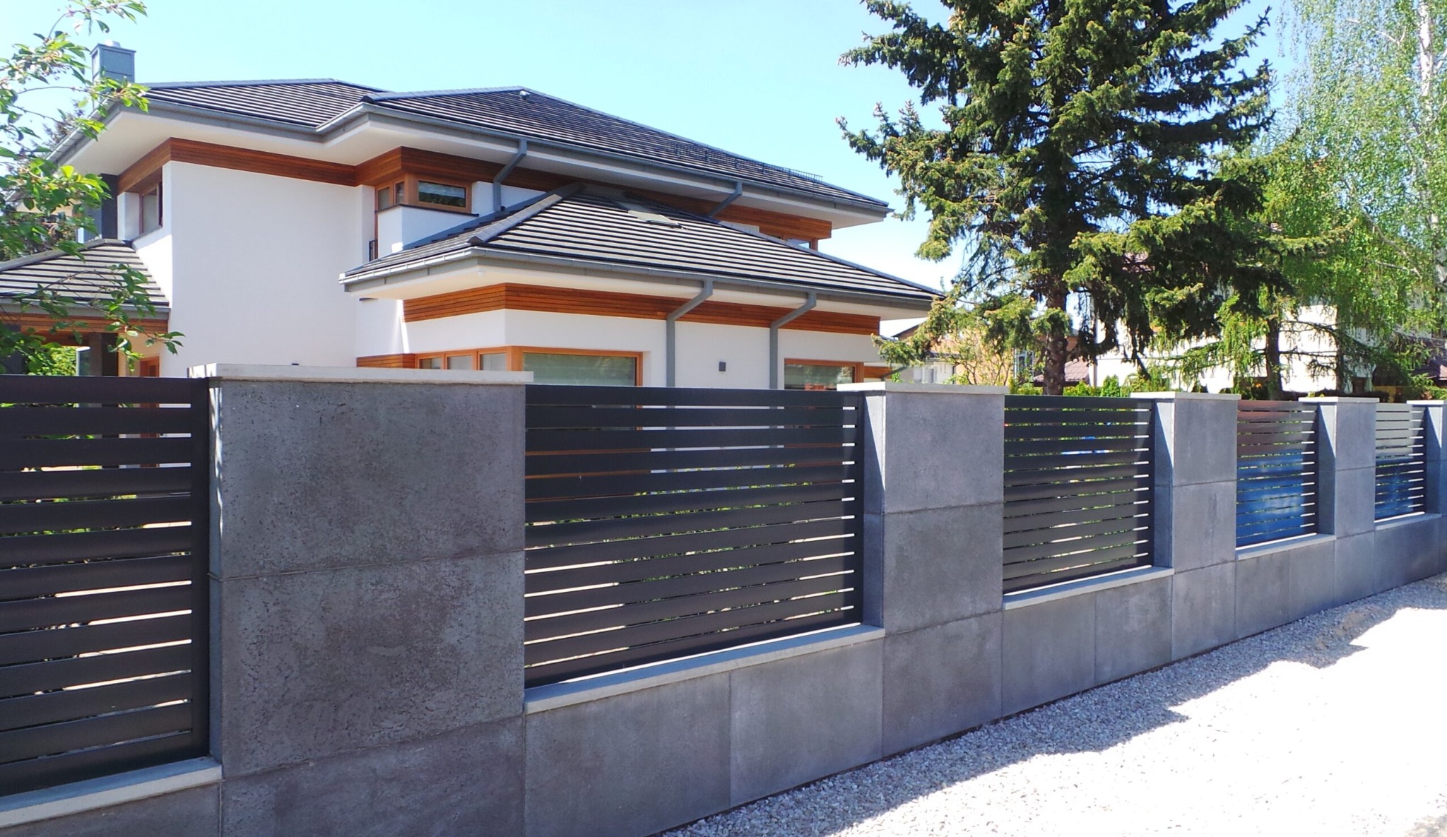 Nowoczesne ogrodzenie z poziomych, ciemnych paneli i betonowych słupków przed domem z białymi ścianami i ciemną dachówką.
