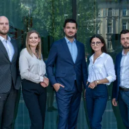 Zespół prawniczy w eleganckich strojach pozuje na tle szklanej fasady budynku, odbijającej architekturę miejską. Profesjonalny wizerunek.