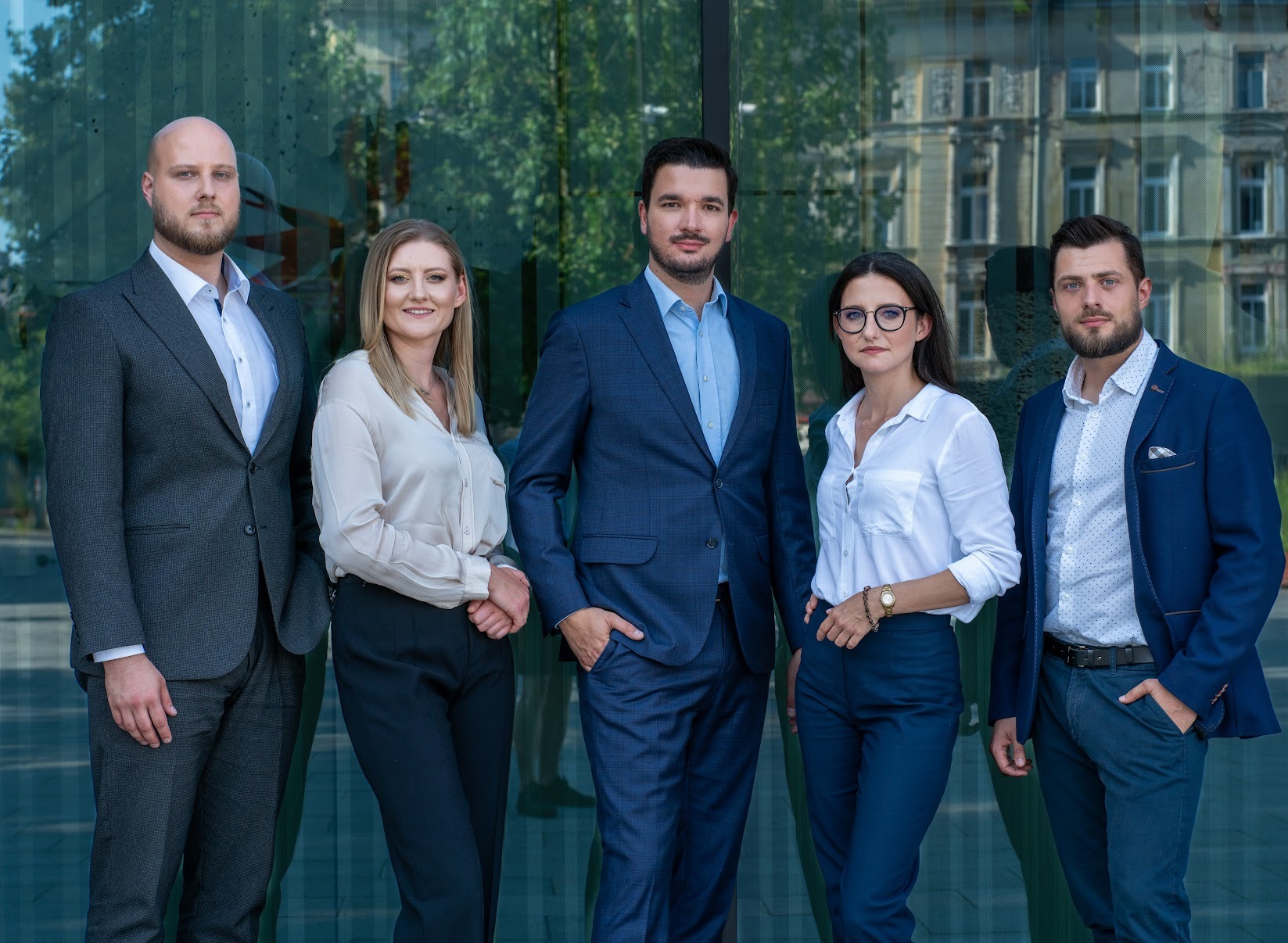 Zespół prawniczy w eleganckich strojach pozuje na tle szklanej fasady budynku, odbijającej architekturę miejską. Profesjonalny wizerunek.
