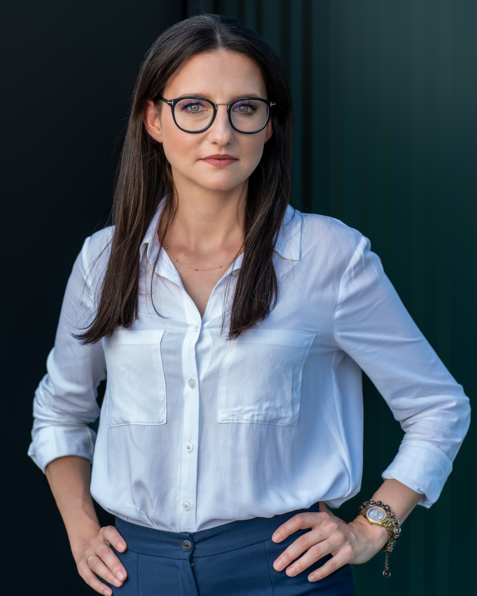 Profesjonalny portret kobiety w okularach, białej koszuli i granatowych spodni na tle ciemnozielonej ściany. Uśmiech, pewny wyraz twarzy, biznesowy wizerunek.