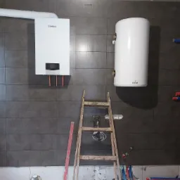 Biały wiszący piec gazowy Vaillant z podłączonymi miedzianymi rurkami i biały bojler Galmet na ścianie z szarych płytek, drabina montażowa i wystające rury instalacyjne w tle.