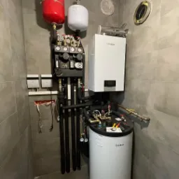 Zestaw urządzeń grzewczych Vaillant w kotłowni wykończonej szarymi płytkami, widoczne zbiorniki, piec, rury, manometry i drobne narzędzia na górze bojlera.