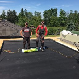 Kompany - Dwóch mężczyzn w roboczych ubraniach stoi na płaskim dachu pokrytym czarną membraną, obok rolki membrany Resitrix SKW i niebieskiego odkurzacza przemysłowego, widoczne narzędzia i skrzynki...