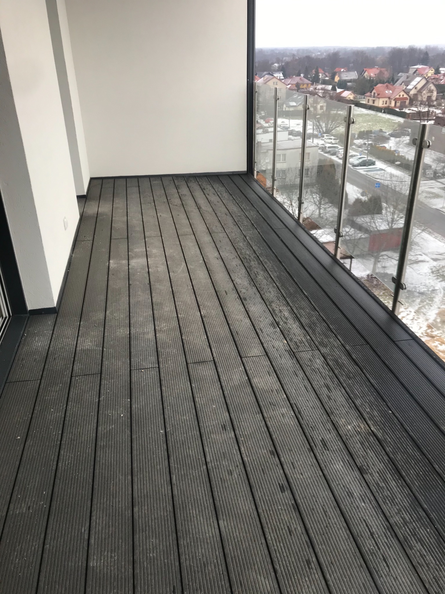 Nowoczesny balkon z szarymi deskami kompozytowymi, szklaną balustradą i widokiem na zimowy krajobraz miejski.